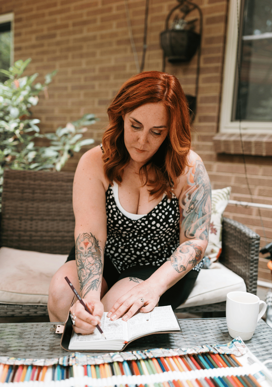 a woman with red hair and tattoos sitting on her porch writing in a journal